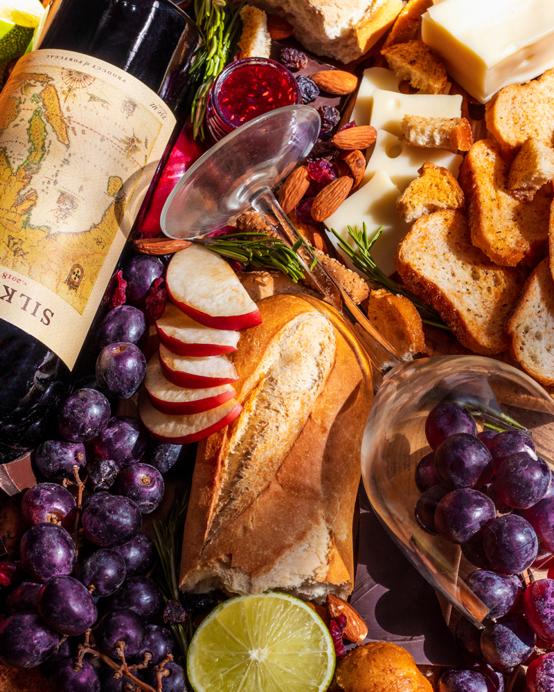 Set design, set styling, commercial photography spec work of fancy red wine. This layout includes the bottle and a wine glass filled with grapes, also surrounded by bread, apple slices, purple grapes, limes, jam, a wine glass, almonds, chocolate, breadsticks, and more.