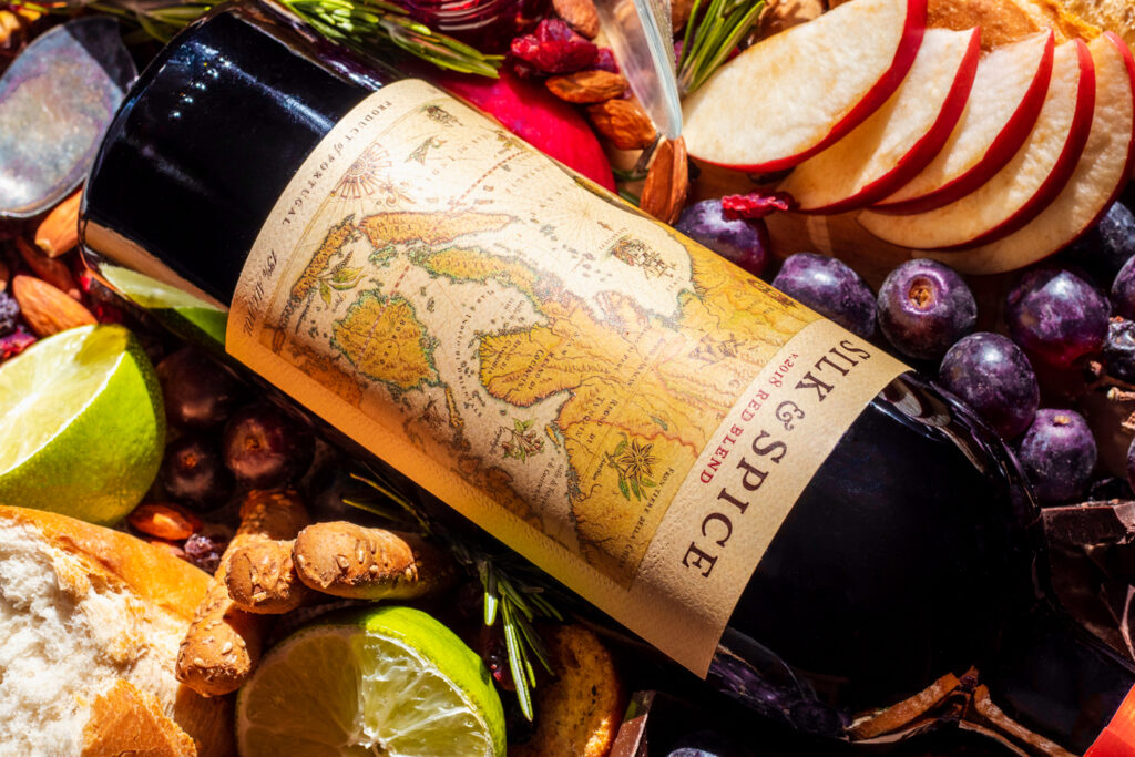 Set design, set styling, commercial photography spec work of fancy red wine. This layout includes the entire bottle surrounded by bread, apple slices, purple grapes, limes, jam, a wine glass, almonds, chocolate, breadsticks, and more