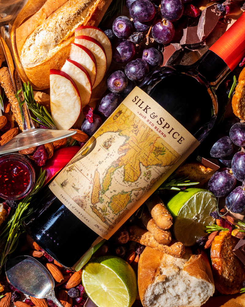 Set design, set styling, commercial photography spec work of fancy red wine. This layout includes the entire bottle surrounded by bread, apple slices, purple grapes, limes, jam, a wine glass, almonds, chocolate, breadsticks, and more.