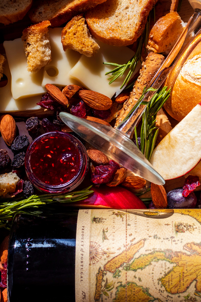 Set design, set styling, commercial photography spec work of fancy red wine. This layout includes the bottle and a wine glass, also surrounded by bread, apple slices, purple grapes, limes, jam, a wine glass, almonds, chocolate, breadsticks, and more.